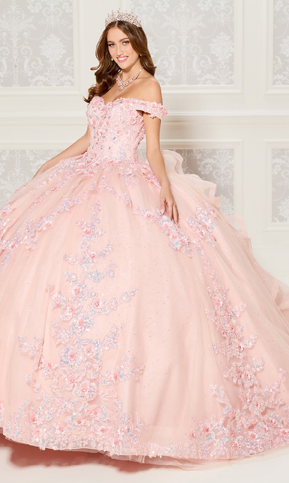 A young woman in the Quinceanera Dress PR30116 By Princesa, featuring pink off-the-shoulder floral details, a tiara, and necklace, stands before an ornate white wall.